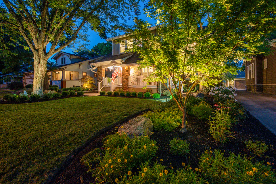 Front landscape with tree uplighting, warm porch lights, lawn, and planted beds at night